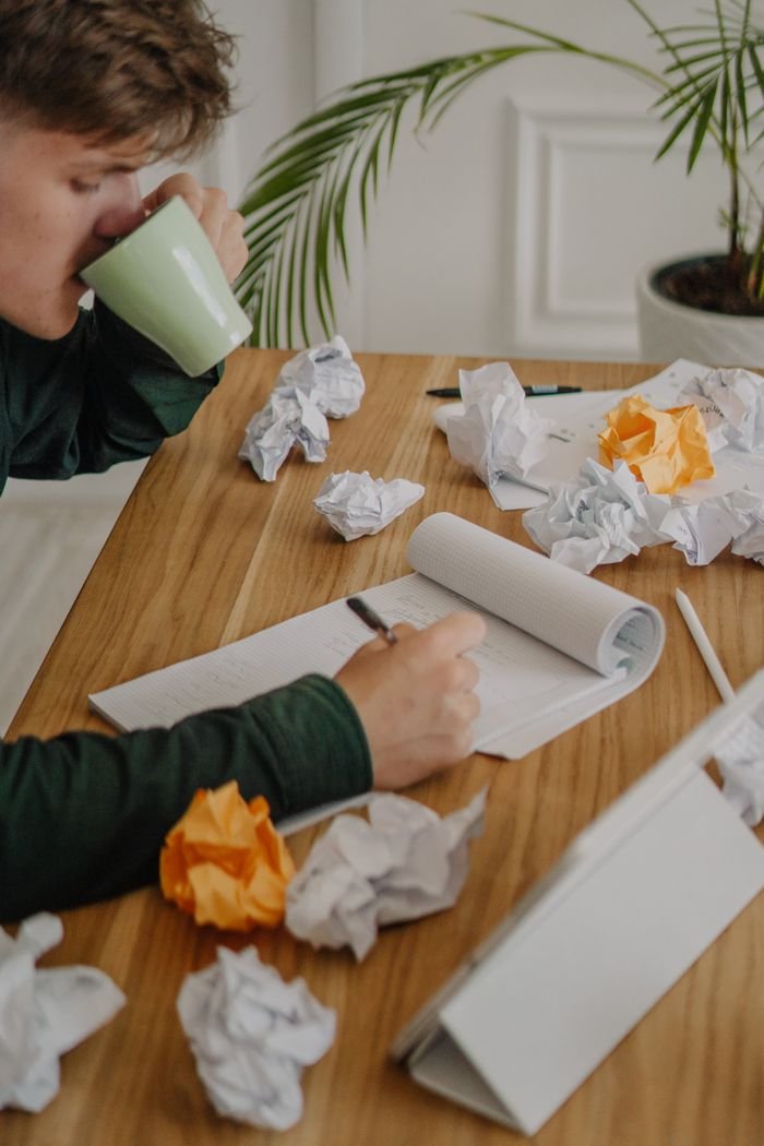 Adult man in green shirt drinking coffee and writing amidst crumpled papers at a wooden desk.